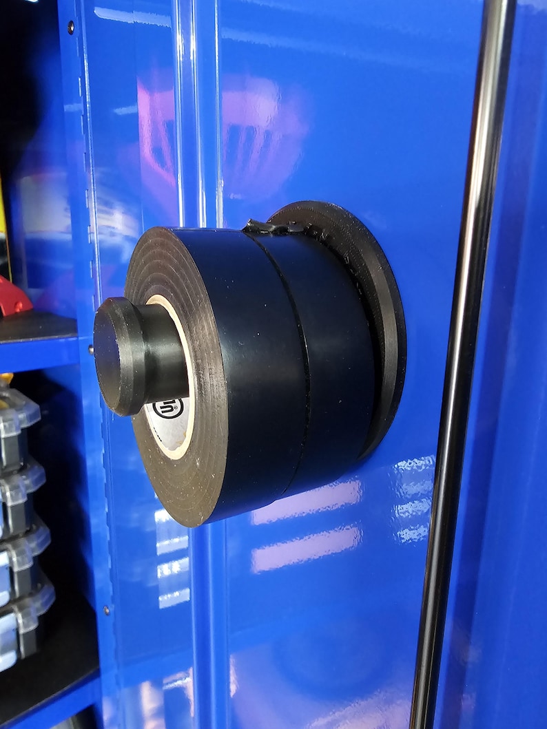 May include: A roll of black electrical tape mounted on a blue surface. The tape is held in place by a black holder. The tape roll has a white label with a logo. A metal rod is visible in the background.