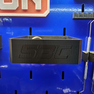 May include: A black, rectangular storage bin with the letters "SBC" embossed on the front. The bin is mounted on a blue pegboard, with other tools and storage accessories visible in the background.