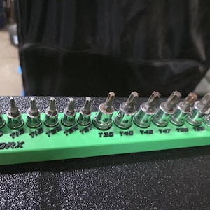May include: A green Torx bit holder with various sizes of Torx bits. The sizes are labeled T9, T10, T15, T20, T25, T27, T30, T40, T45, T47, T50, and T55. The holder is on a textured surface.