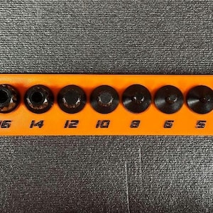 May include: An orange tool holder with black sockets of varying sizes, labeled from 4 to 18. The sockets are arranged in a row, with the size numbers printed in white. The brand name is printed in orange on the right side.