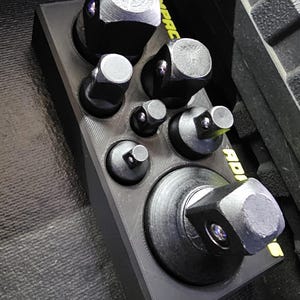 May include: A black socket set with various sized sockets arranged in a black holder. The sockets are made of metal and have a square shape. The holder is rectangular and has a yellow label with the word "Impact" on it.