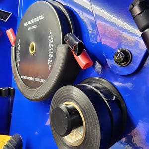 May include: Black and gray electrical tape and a black and gray Maddox retractable test lead on a blue surface. The test lead has red and black caps. The text on the test lead reads "MADDOX SKU:58024 TO REWIND-HOLD TOP & ROTATE BOTTOM".
