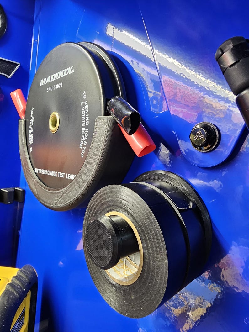 May include: Black and gray electrical tape and a black and gray Maddox retractable test lead on a blue surface. The test lead has red and black caps. The text on the test lead reads "MADDOX SKU:58024 TO REWIND-HOLD TOP & ROTATE BOTTOM".