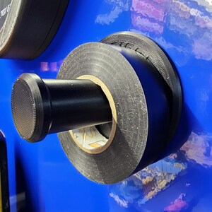 May include: Close-up of a black electrical tape roll mounted on a blue surface. The tape is wrapped around a cardboard core, with a black cylindrical handle. The background is a glossy blue.
