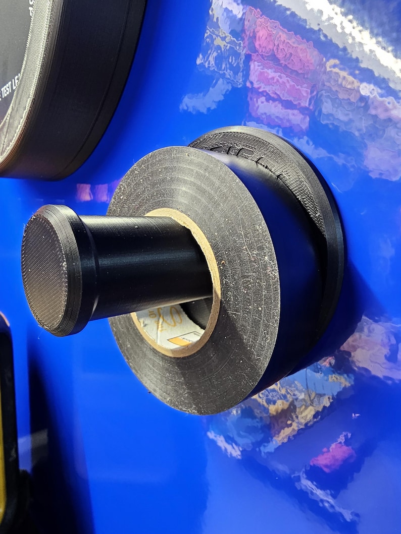 May include: Close-up of a black electrical tape roll mounted on a blue surface. The tape is wrapped around a cardboard core, with a black cylindrical handle. The background is a glossy blue.