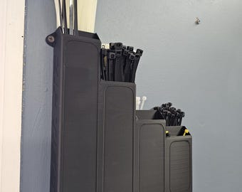 May include: A black, tiered cable tie organizer mounted on a wall. The organizer holds various sizes of cable ties in white and black. The organizer is attached to the wall with screws and is designed to keep cable ties organized and accessible.