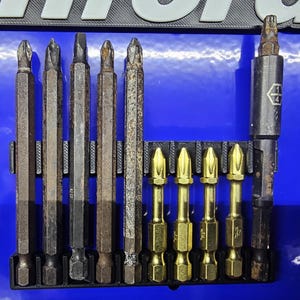 May include: A set of various screwdriver bits in a black holder. The bits are a mix of silver, gold, and brown, showing signs of use. The holder is mounted on a blue surface.
