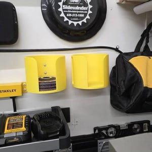 Puede incluir: Una escena de taller con un contenedor de almacenamiento amarillo que contiene hojas de sierra circular. Una bolsa de herramientas DeWalt negra y amarilla cuelga cerca. También se ven un nivel Stanley y un cargador DeWalt.