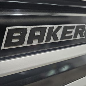 May include: A black and silver emblem featuring the word BAKER in bold, raised black capital letters set against a textured silver rectangular background, mounted on a black surface with horizontal silver trim accents below the emblem.