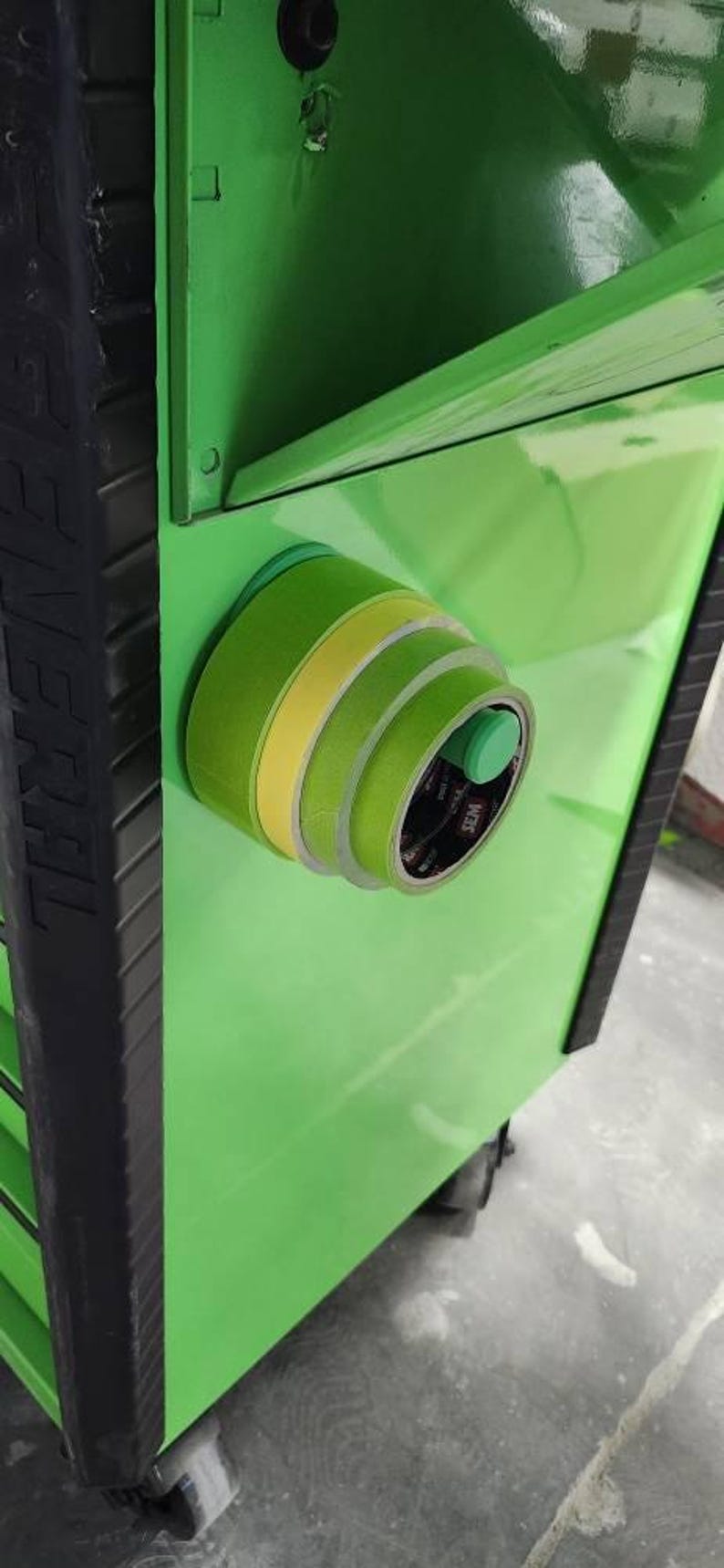 May include: A close-up of a green tool cabinet with rolls of green and yellow masking tape attached to the side. The tape is held in place by a magnetic holder. The cabinet is a bright, glossy green.