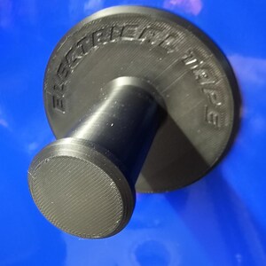 May include: A black, 3D-printed object with the text "ELECTRIC WHEEL" embossed on a circular top. The object has a cylindrical stem and a rounded base, all in black. The background is a solid blue.