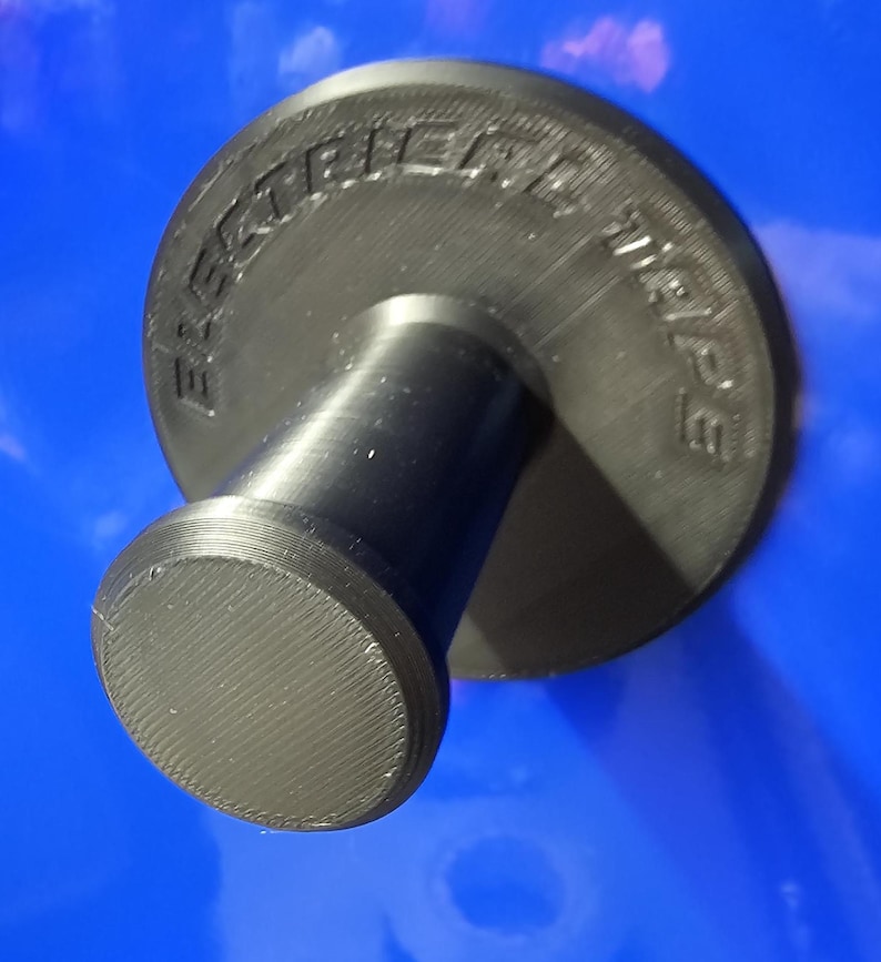 May include: A black, 3D-printed object with the text "ELECTRIC WHEEL" embossed on a circular top. The object has a cylindrical stem and a rounded base, all in black. The background is a solid blue.