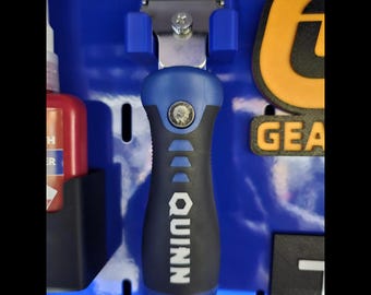 May include: A Quinn brand scraper tool with a blue and black ergonomic handle, featuring a metal blade head secured by a silver screw, mounted vertically on a blue pegboard panel alongside a red bottle and a yellow and black gear logo.