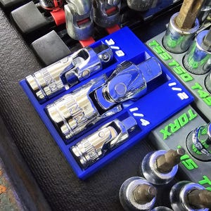 May include: A blue organizer tray holding three chrome universal joint sockets labeled 1/4, 1/2, and 3/8. Other sockets and tools are visible in the background. The image is well-lit and shows the tools in detail.