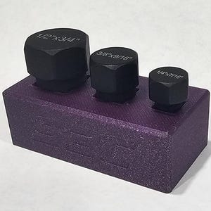 May include: A purple tool holder with three black hex nuts of varying sizes. The largest nut is labeled "1/2"x3/4", the middle one "3/8"x9/16", and the smallest "1/4"x7/16". The holder is made of a textured material.