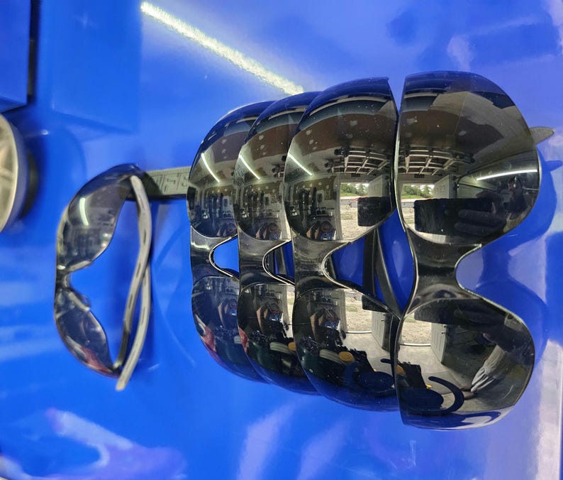 May include: A display of several pairs of mirrored sunglasses with a unique, curved design. The sunglasses are arranged on a blue surface, reflecting the surrounding environment. The frames are a shiny, metallic color.