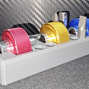 May include: A gray tool organizer holding various colored bits. The bits include red, gold, and blue, with silver and black accents. The organizer has the letters "SSC" on the side and is designed to hold multiple tools.