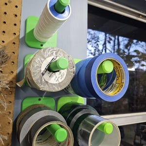 May include: A wall-mounted tape dispenser system with several rolls of tape in various colors and sizes. The system is made of green plastic and holds rolls of tape with white, blue, gray, and black colors. The tape rolls are mounted on green pegs.