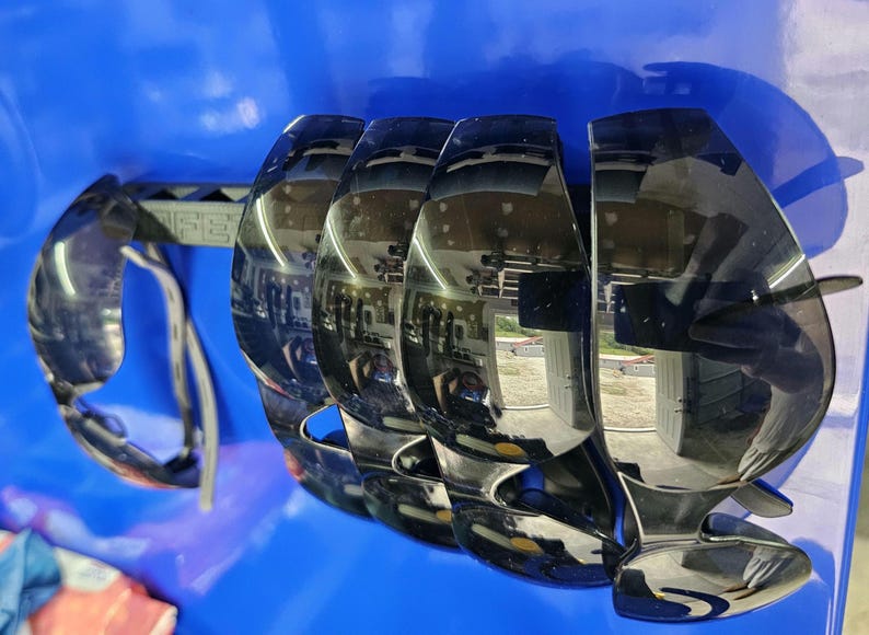May include: Black sunglasses with a reflective surface. The sunglasses have a unique, curved design with a thick frame and a bridge. The lenses are dark, and the arms are thin and gray. The sunglasses are set against a blue background.