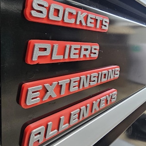 May include: Close-up of four red and silver tool labels: "Sockets", "Pliers", "Extensions", and "Allen Keys". The labels are mounted on a black surface, with a silver trim visible.