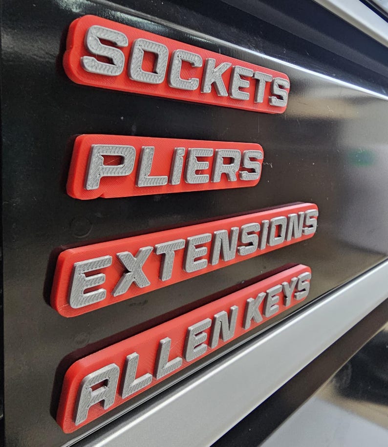 May include: Close-up of four red and silver tool labels: "Sockets", "Pliers", "Extensions", and "Allen Keys". The labels are mounted on a black surface, with a silver trim visible.