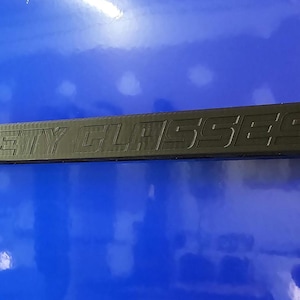 May include: A black safety glasses frame with the words "SAFETY GLASSES" embossed on the top. The frame is rectangular and appears to be made of a durable material. The background is a glossy blue surface.