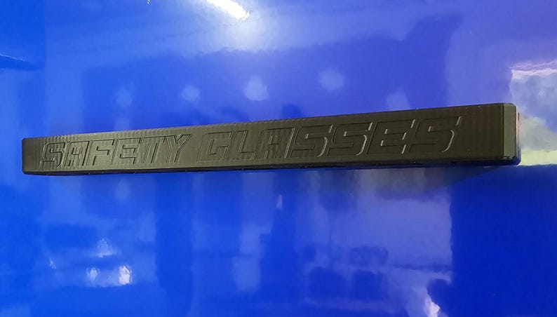 May include: A black safety glasses frame with the words "SAFETY GLASSES" embossed on the top. The frame is rectangular and appears to be made of a durable material. The background is a glossy blue surface.