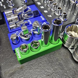 May include: A collection of chrome sockets and ratchets arranged on blue and green organizers. The sockets are various sizes and shapes, designed for mechanical work. The image is well-lit, showcasing the tools' reflective surfaces.