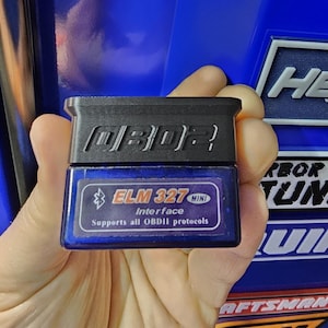 May include: A black and blue OBD2 ELM327 Mini Interface device. The device has the text "OBD2" on the top and "ELM 327 MINI Interface Supports all OBDII protocols" on the bottom. The device is held in a hand.