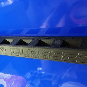 May include: Black rectangular safety glasses holder with the words "SAFETY GLASSES" embossed on the front. The holder has multiple compartments for storing eyewear. The holder is made of a dark material and sits on a blue surface.