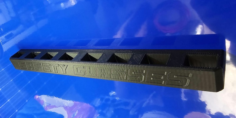 May include: Black rectangular safety glasses holder with the words "SAFETY GLASSES" embossed on the front. The holder has multiple compartments for storing eyewear. The holder is made of a dark material and sits on a blue surface.
