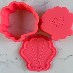 May include: Three pink plastic cookie cutters shaped like seashells. The largest cutter is a simple seashell shape, the middle cutter is a seashell with a raised design, and the smallest cutter is a seashell with a raised design and a small circle in the center.
