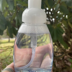 May include: A clear plastic bottle with a white pump dispenser filled with a clear liquid. The bottle is in a hand.