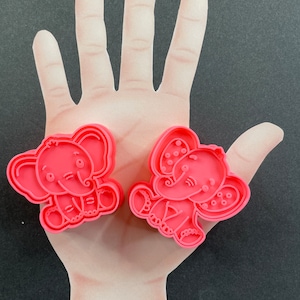 May include: Two pink plastic cookie cutters shaped like elephants. The elephants are sitting with their trunks raised and have detailed features.