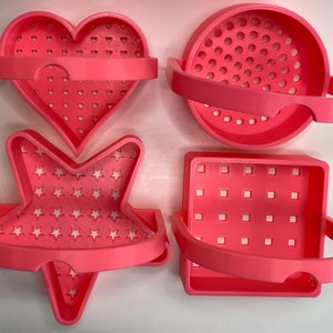 May include: Four pink plastic soap dishes with different shapes: a heart, a star, a square, and a circle. Each dish has a handle and a patterned design.