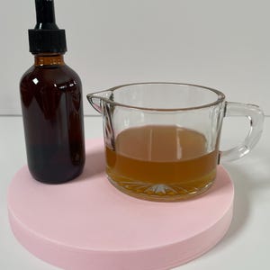 May include: A brown glass bottle with a dropper top and a clear glass measuring cup filled with a light brown liquid, both sitting on a pink circular surface.