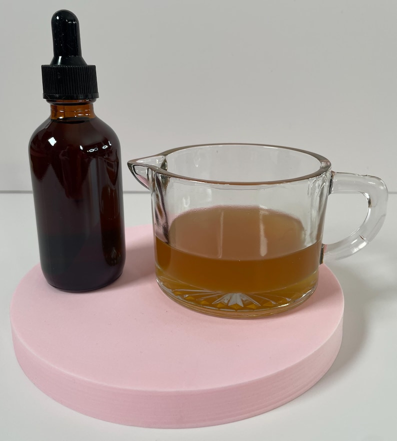May include: A brown glass bottle with a dropper top and a clear glass measuring cup filled with a light brown liquid, both sitting on a pink circular surface.