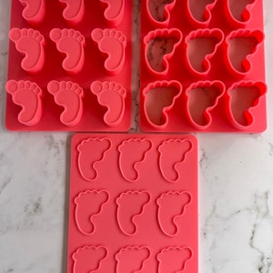 May include: Three pink silicone molds for making baby foot-shaped treats. Each mold has multiple cavities, with the largest mold having nine cavities.