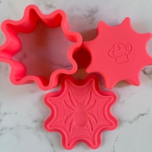 May include: Three pink plastic cookie cutters shaped like stars with a spider web and spider design, a star with a chef hat design, and a plain star. The star with the chef hat has the letters "TSC" inside the hat.