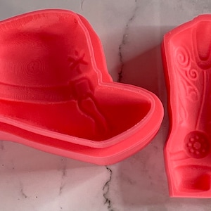 May include: Two pink silicone molds for baking, one shaped like a cowboy hat and the other shaped like a cowboy boot.