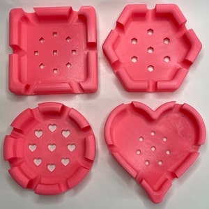 May include: Four pink silicone molds with different shapes: a square, a hexagon, a heart, and a circle. Each mold has multiple small holes for making soap, resin, or other crafts.