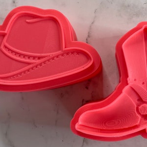 May include: Two pink plastic cookie cutters, one shaped like a cowboy hat and the other shaped like a cowboy boot.