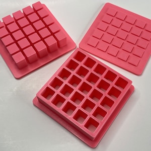 May include: Three pink plastic molds for making square-shaped objects. Each mold has a grid of squares, with the top mold having raised squares and the bottom two molds having indented squares.