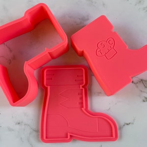 May include: Three pink plastic cookie cutters shaped like boots. One boot has a chef hat design on the top.