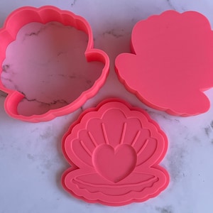 May include: Three pink plastic cookie cutters shaped like seashells. The largest cutter is a simple seashell shape, the medium cutter is a seashell with a heart inside, and the smallest cutter is a seashell with a detailed design.