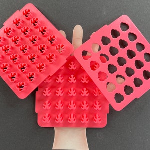May include: Three pink plastic molds for making fondant or gum paste decorations. The molds feature different designs, including leaves, flowers, and a cloud-like shape.