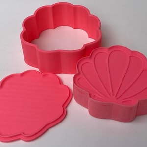 May include: A pink seashell-shaped trinket box with a removable lid and a separate base. The box has a detailed seashell design on the lid and base.