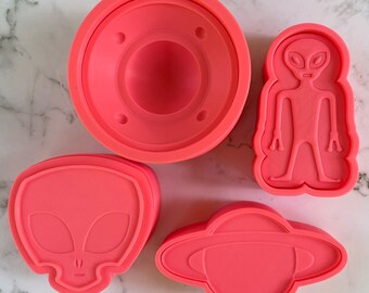 Op de afbeelding: Vier roze siliconen mallen voor het bakken, gevormd als een alienhoofd, een alienlichaam, een vliegende schotel en een ronde vorm met een verhoogd midden.