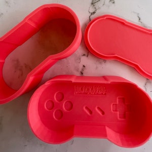 May include: Three coral-colored cookie cutters in the shape of a classic video game controller. The set includes the controller base, a top piece, and a long, oval-shaped piece. The controller base has the words "SUPER NINTENDO" printed on it.