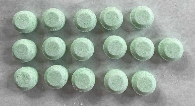May include: A close-up view of fifteen round, green bath bombs arranged in three rows of five on a white surface.